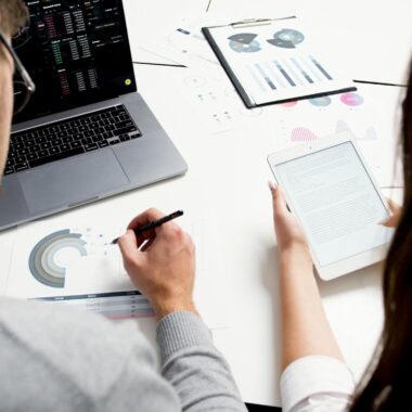 Two business professionals analyzing financial reports using a laptop and tablet in a modern office setting.